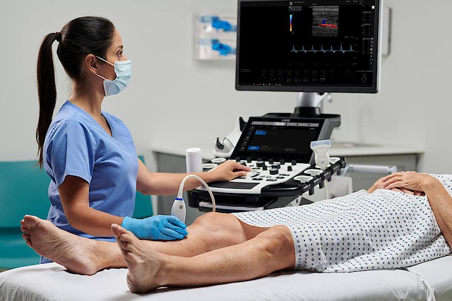 Imaginea prezintă un medic ce efectuează un examen ecografic vascular al unui pacient.